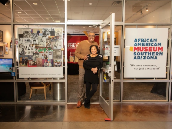 A smiling couple stands in the doorway of the African American Museum of Southern Arizona, welcoming visitors. The woman, dressed in black, leans slightly against the open glass door, while the man, wearing a brown sweater, stands behind her with his arm around her. The museum entrance features a large sign that reads, 'African American Museum of Southern Arizona' with the tagline, 'We are a movement, not just a museum!' A collage of historical African American figures and moments is displayed on the glass