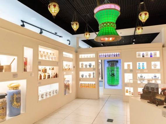 The interior of the Coit Museum of Pharmacy and Health Sciences, showcasing a collection of historical pharmacy artifacts. The walls are lined with illuminated display cases containing antique medicine bottles, ornate apothecary jars, and pharmaceutical tools. A large, green vintage-style glass sign hangs from the ceiling, reading 'Company Founded 1886.' Stained-glass lanterns with intricate designs provide additional ambiance. In the background, a digital display features a glowing green bottle