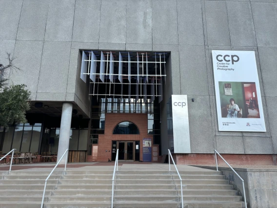 The exterior of the Center for Creative Photography (CCP) at the University of Arizona, featuring a modern architectural design with a mix of concrete, brick, and glass elements. A large banner on the right side of the building displays the CCP logo along with an image of a woman sitting in a vintage-style room, promoting an exhibition. The entrance has a set of stairs leading up to glass doors, framed by an arched brick design. Above the entrance, a unique structure with angled panels provides a dynamic vi