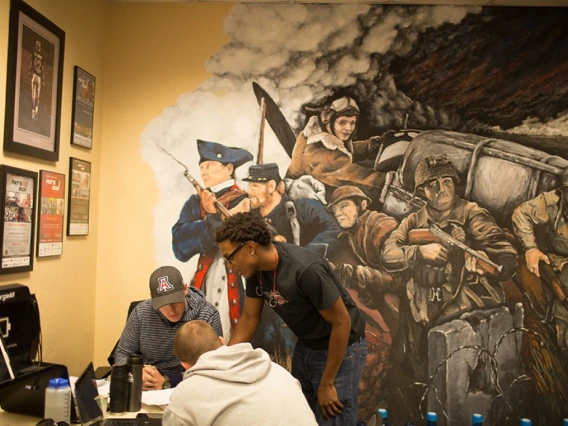 three people working at a desk in front of a military mural painted on the wall
