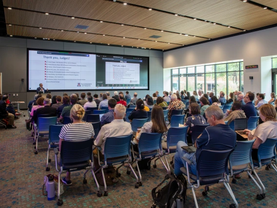 A room full of people sitting in chairs listening to a man standing at a podium in front of two large screens.