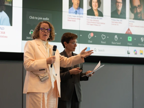 Two individuals, one holding a microphone and the other holding a script, speak in front of a large screen.