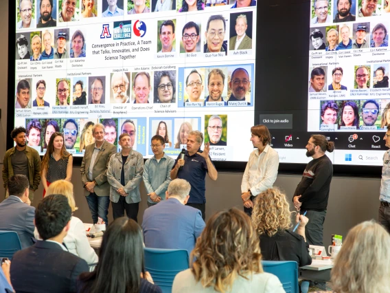 A diverse group of individuals stand in front of two large screens showing photos of additional team members.
