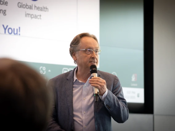 A man in glasses holding a microphone speaks in front of a large screen.