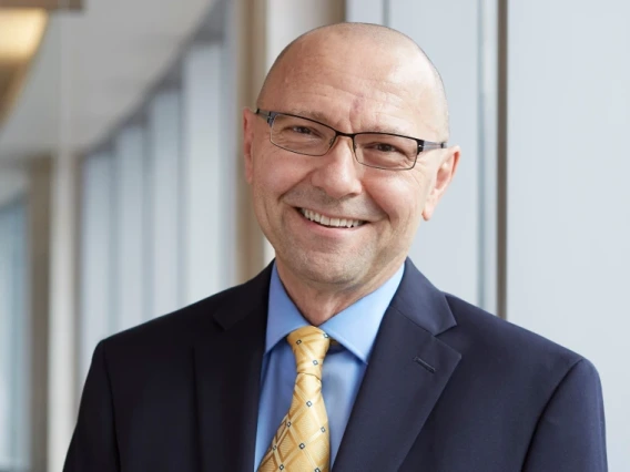 A photograph of Dan Theodorescu, smiling, wearing rectangular glasses and a navy suit with a yellow tie.