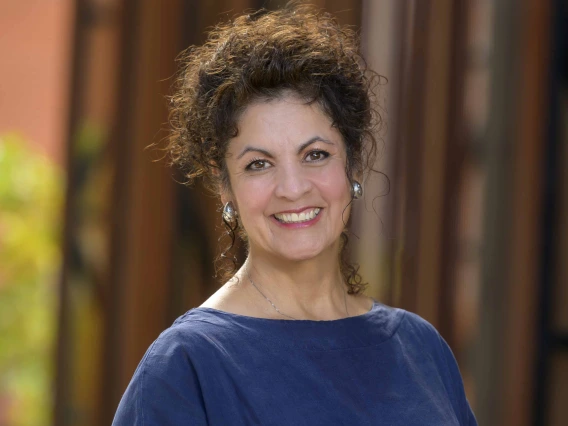 Smiling woman with dark curly hair and silver earrings, wearing a navy blouse, stands outdoors in soft focus architectural background with warm light and blurred vertical elements.