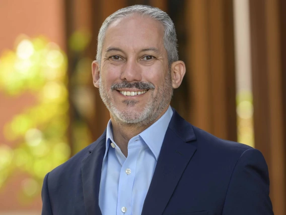 Smiling man with short gray hair and beard, wearing a navy blazer and light blue shirt, stands in front of a softly blurred background with warm tones and greenery.