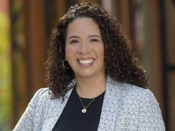 Smiling woman with long curly hair, wearing a light gray blazer, black top, and gold jewelry, stands in front of a softly blurred architectural background.