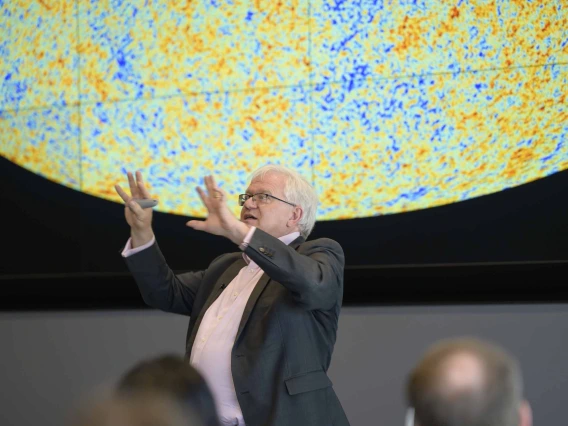 A man with white hair holds his hands in the air as he presents to an audience with a large circular projection of yellow, orange and blue dots behind him. 