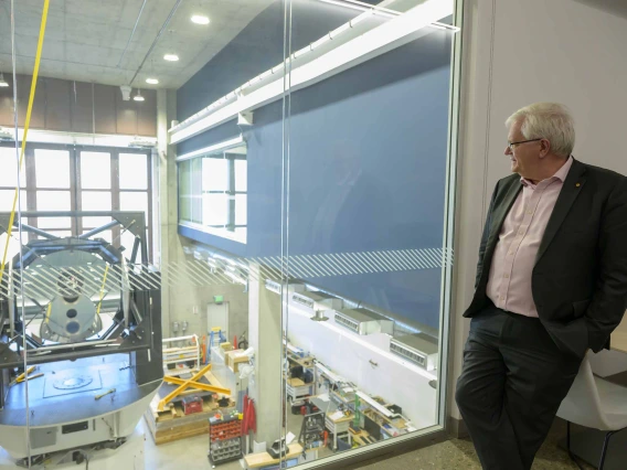 A man with white hair leans against a desk while looking through a window at a large machine in laboratory-like workspace. 