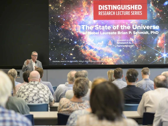 View from the back of a lecture hall over the heads of the audience with a projection of space with stars and text that reads, “Distinguished Research Lecture Series”. 