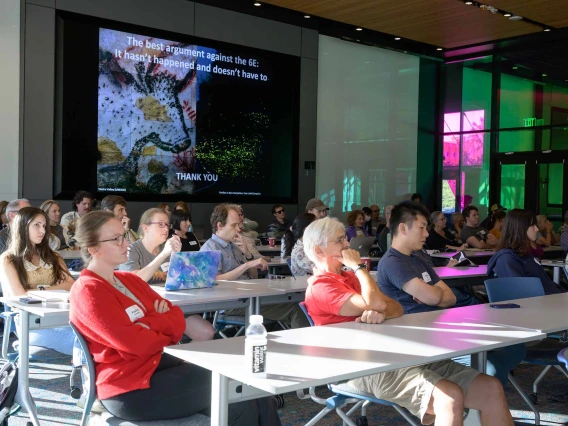 An audience sits as tables listening to a lecture as a presentation is projected on a screen. 