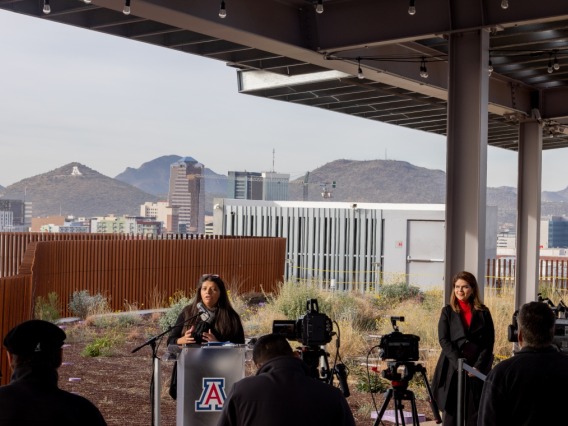 A woman speaks behind a podium with the UArizona logo in front of a backdrop of the Catalina Mountain range