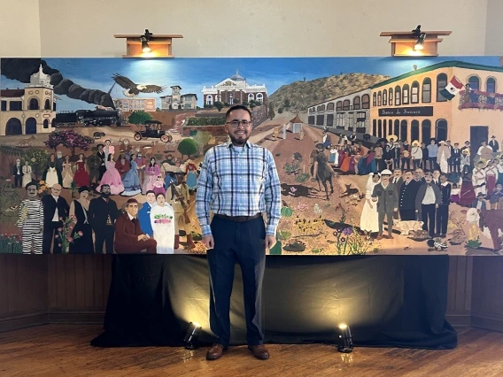 A man stands smiling in front of a large, colorful mural that depicts a historical and cultural scene with various figures, buildings, animals, and landscapes, likely representing a community or region's heritage. The artwork includes people in traditional and historical attire, with a Mexican flag visible on one of the buildings.