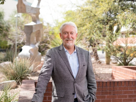 A man with white hair and a beard stands outdoors, leaning against a brick wall with one hand in his pocket. He is wearing a light blue dress shirt and a gray blazer, smiling at the camera.