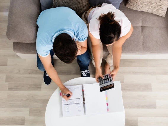 Overhead view of a man and woman sitting on a sofa, reviewing financial documents. The man is writing on a piece of paper in a binder while the woman uses a calculator, both appearing focused on their task.