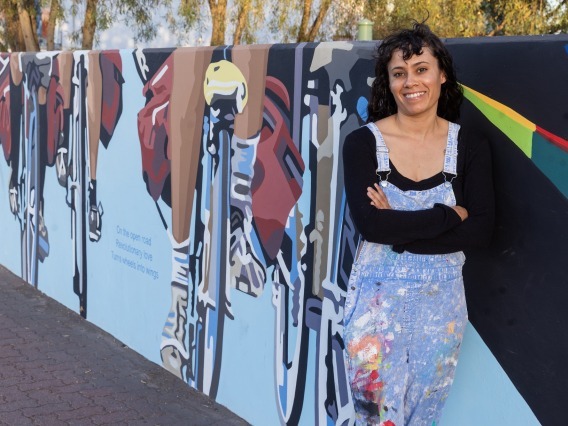A woman in a black, long sleeve shirt and paint covered overalls leans against a mural painted on a wall.