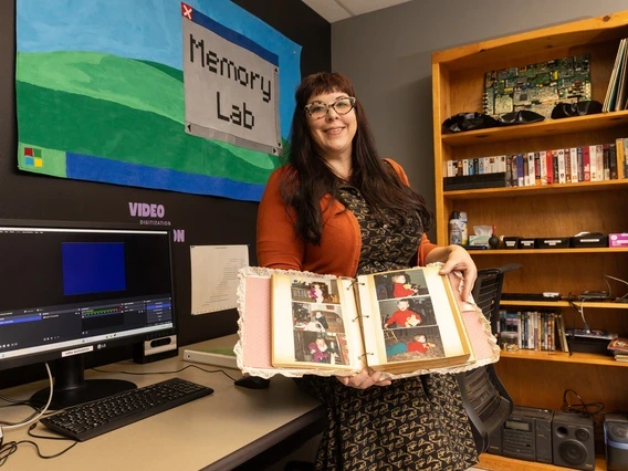 A woman with long dark hair and glasses stands in a digital preservation lab, holding open a scrapbook filled with old family photos. She wears a patterned dress with an orange cardigan and smiles at the camera. Behind her, a colorful banner reading 'Memory Lab' hangs on the wall, with a workstation featuring a computer, VHS tapes, and archival equipment visible.