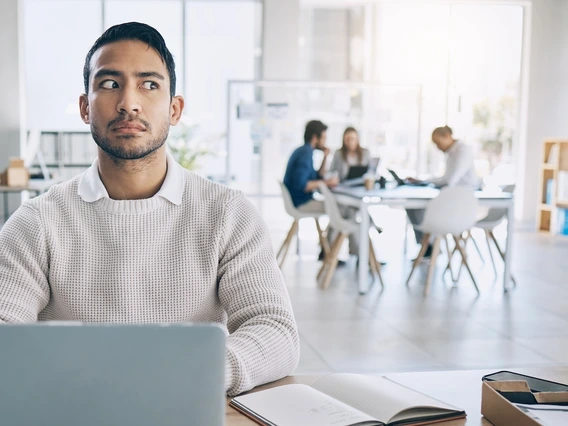 A man sitting at a desk with a laptop and open notebook in front of him looks suspiciously to the side. He is wearing a white knit sweater over a collared shirt. In the background, a group of four people are having a meeting at a table in a brightly lit, modern office space.