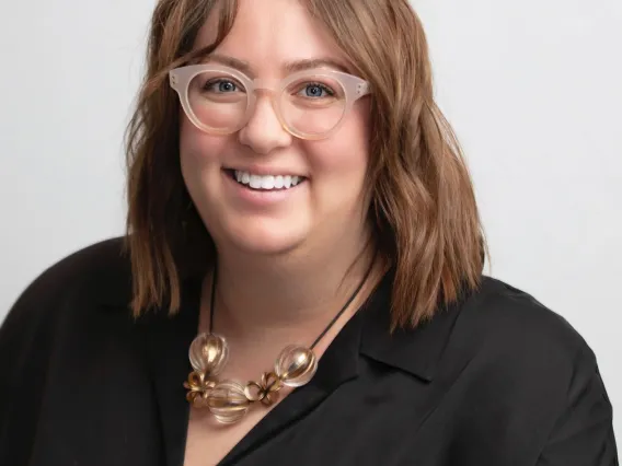 Photograph of Emily Litvack in a black top, chunky necklace, and semi-opaque white glasses