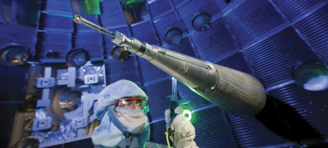 A technician in full cleanroom attire, including a face shield and hood, stands near a long, conical instrument used to position fuel targets inside the National Ignition Facility. The background shows the interior of the target chamber, with glowing blue lighting and metal structures creating a high-tech atmosphere.