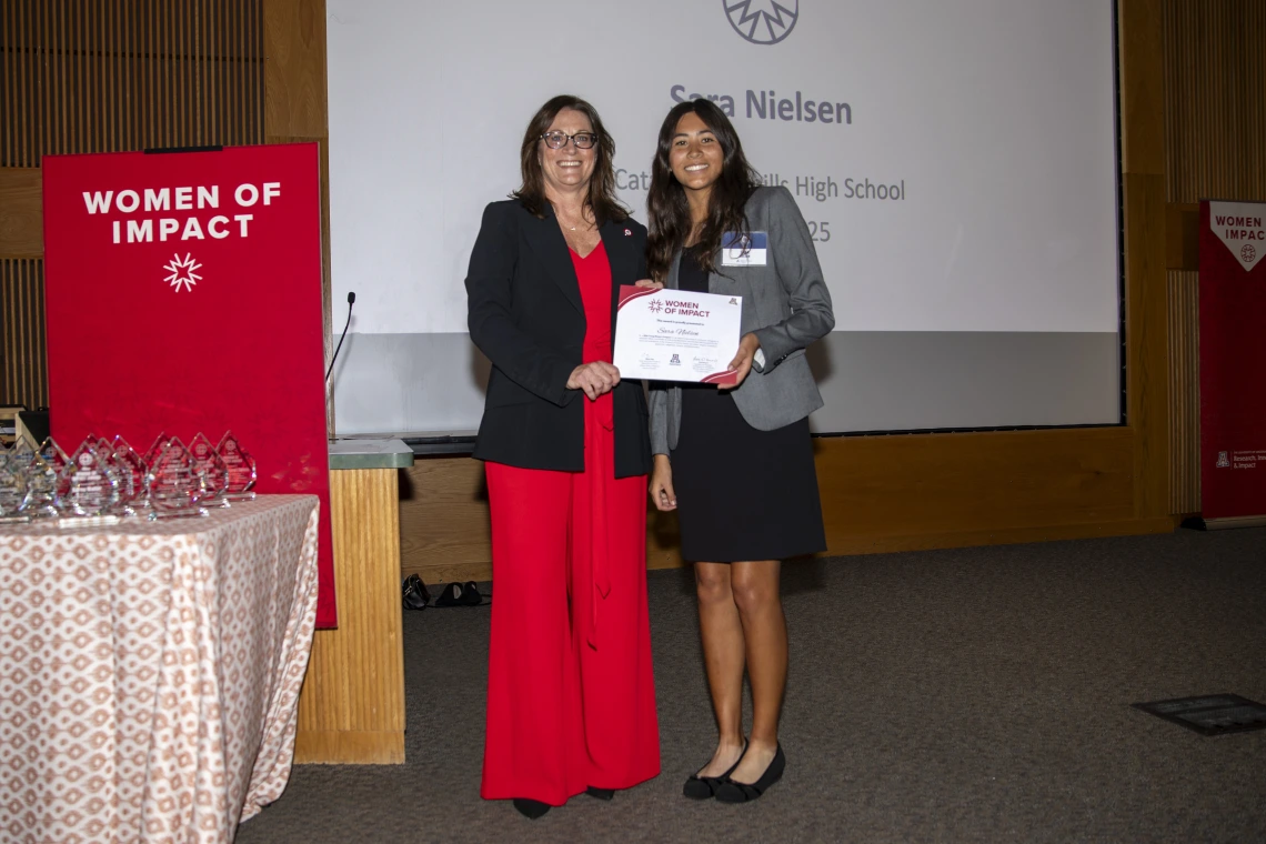 Two women holding a certificate pose for a photo in front of a projector screen.