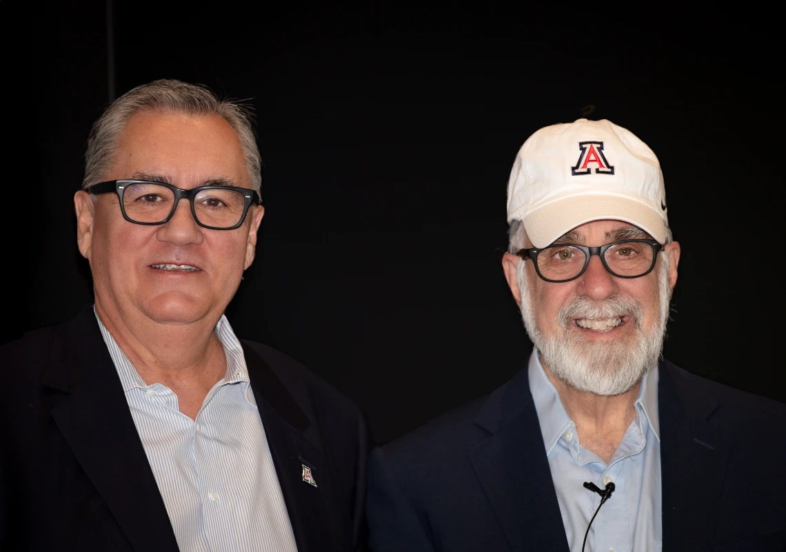 Two men in glasses pose for a picture in front of a black backdrop.