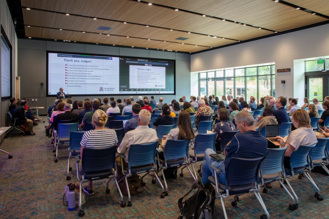 A room full of people sitting in chairs listening to a man standing at a podium in front of two large screens.