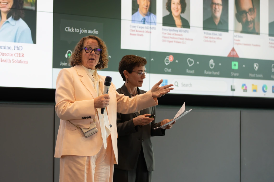 Two individuals, one holding a microphone and the other holding a script, speak in front of a large screen.