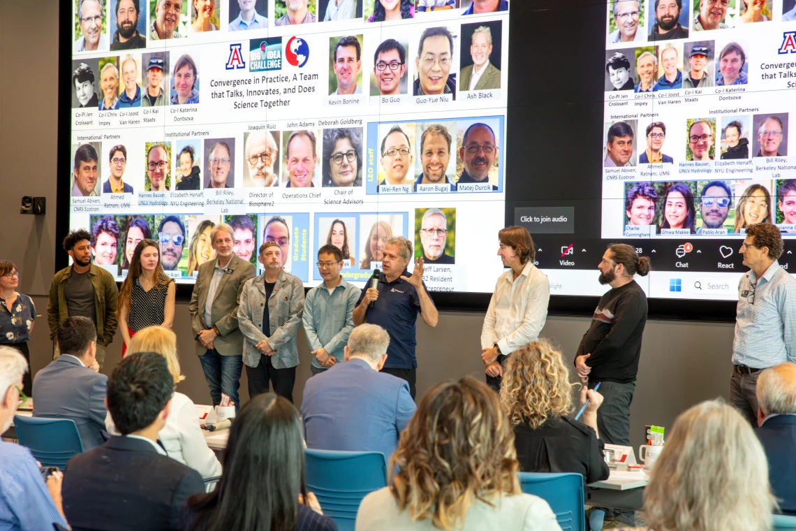A diverse group of individuals stand in front of two large screens showing photos of additional team members.
