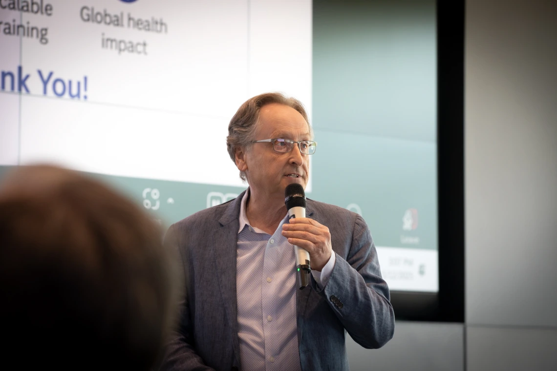 A man in glasses holding a microphone speaks in front of a large screen.
