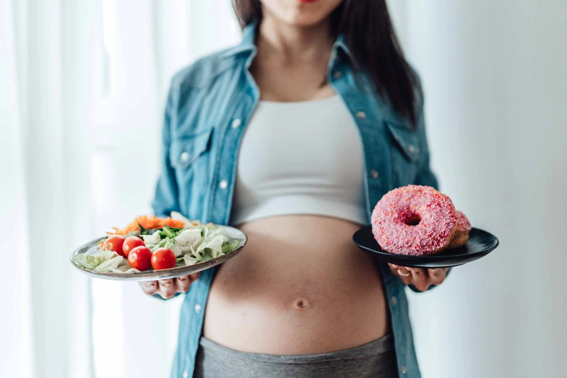 A pregnant woman stands with one plate of salad and another with a donut