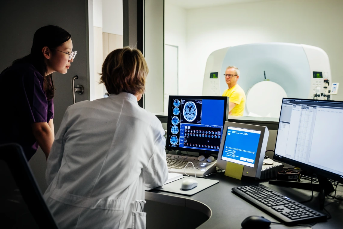 A female doctor and a nurse observing medical imaging results on a computer screen in a hospital, while a patient undergoes an imaging procedure. They are located in a control room, with screens displaying medical data and imaging results.
