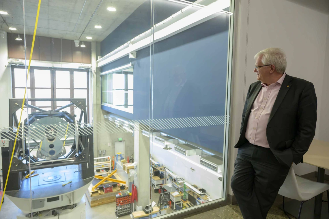 A man with white hair leans against a desk while looking through a window at a large machine in laboratory-like workspace. 
