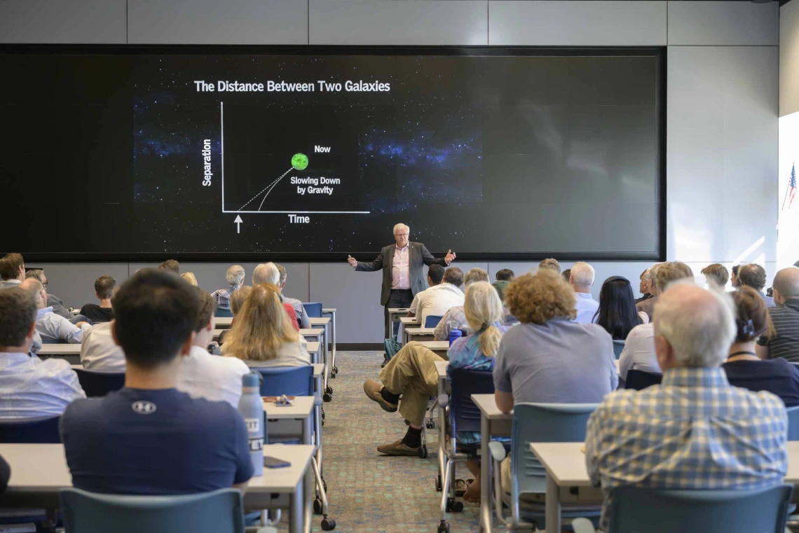 A man with white hair stands in front of an audience talking about a line chart on the screen behind him. 