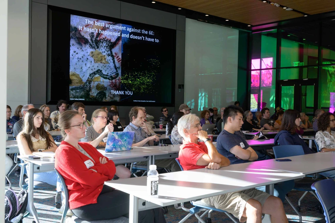 An audience sits as tables listening to a lecture as a presentation is projected on a screen. 