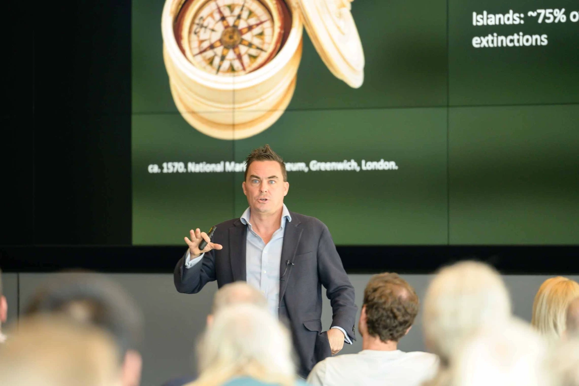 A man stands in front of an audience giving a presentation with a projection on the screen behind him. 