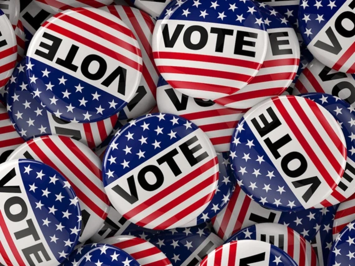 A pile of red, white, and blue "VOTE" buttons featuring stars and stripes, designed to resemble the American flag.