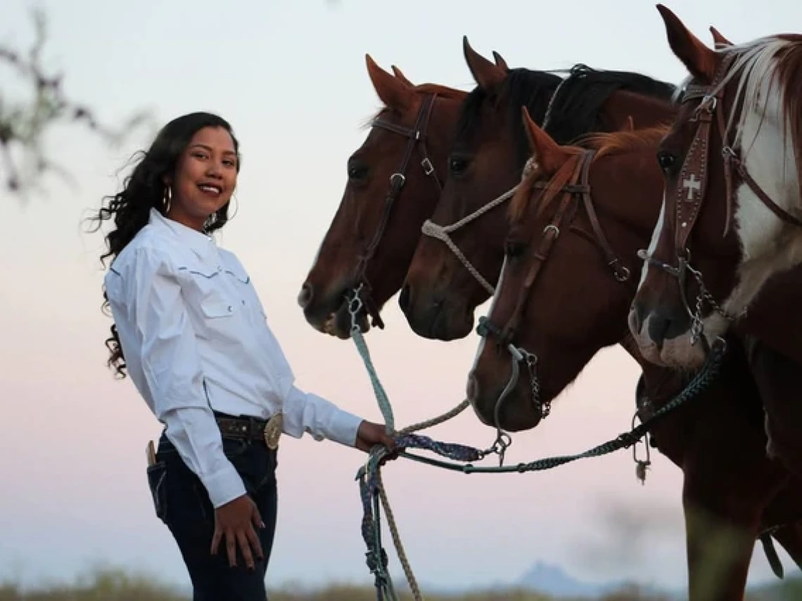 A woman with long dark hair wearing dark denim jeans and a long sleeved white formal shirt stands in front of four horses in an outdoor environment. The sun is setting in the background. The woman is holding leads to all four horses.