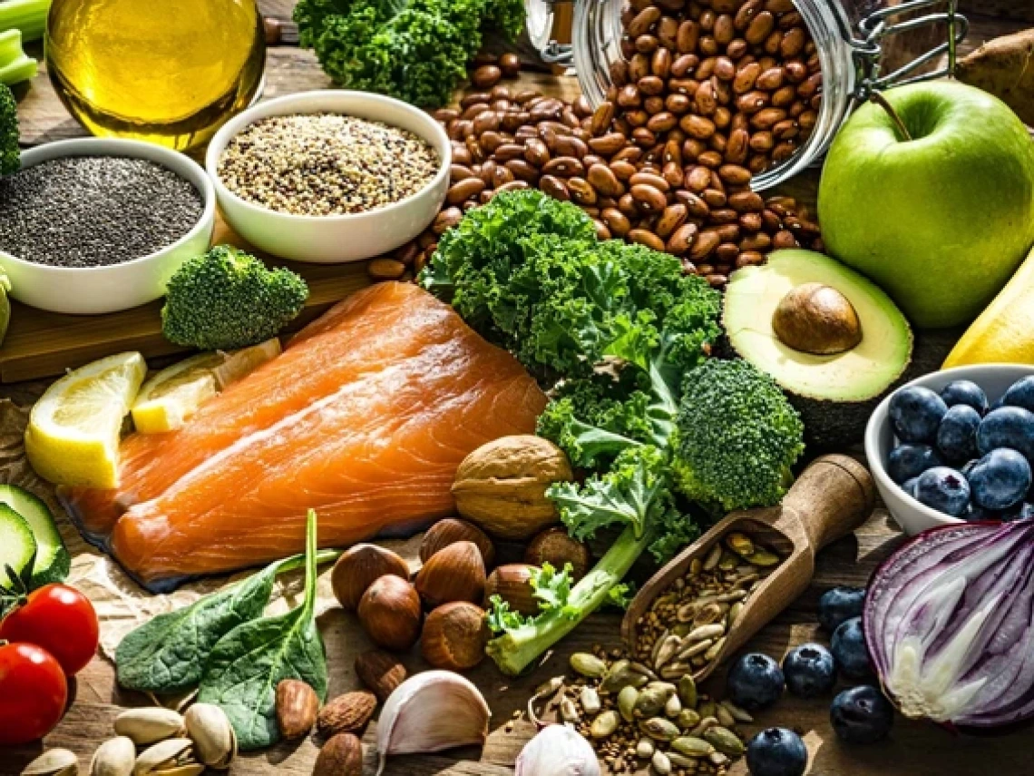 Colorful spread of healthy whole foods including a raw salmon fillet, avocado, blueberries, apple, banana, kale, broccoli, cherry tomatoes, red onion, lemon, chia seeds, quinoa, beans, assorted nuts and seeds, olive oil, and various leafy greens arranged on a rustic wooden surface. A glass jar spills over with pinto beans in the background, adding texture and warmth to the nutrient-rich display.