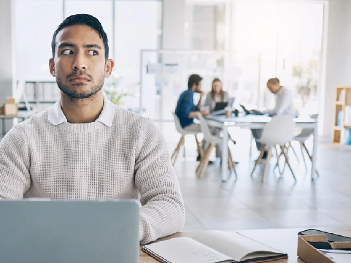 A man sitting at a desk with a laptop and open notebook in front of him looks suspiciously to the side. He is wearing a white knit sweater over a collared shirt. In the background, a group of four people are having a meeting at a table in a brightly lit, modern office space.