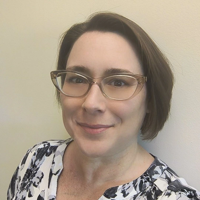 A headshot of a caucasian woman with pale skin, a straight brunette bob, and glasses wearing a black and white flowered shirt