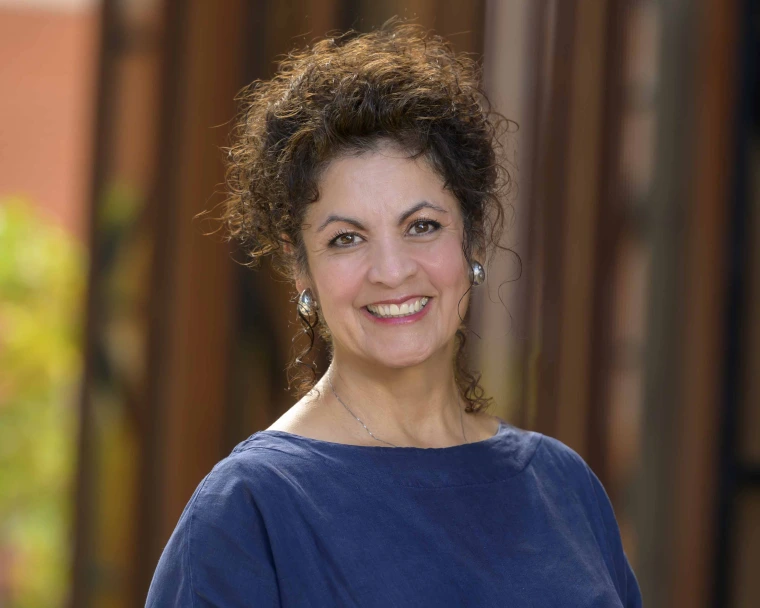 Smiling woman with dark curly hair and silver earrings, wearing a navy blouse, stands outdoors in soft focus architectural background with warm light and blurred vertical elements.