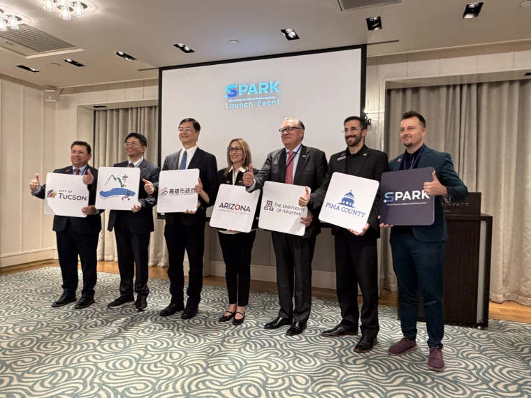 Seven people pose in front of a screen. Each holds a sign indicating the organization they represent.