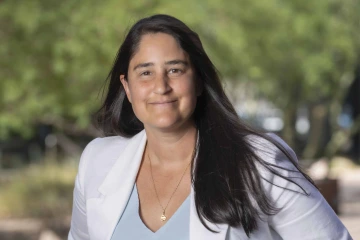 Portrait of Maya Kaelberer wearing a white blazer in an outdoor setting with trees in the background