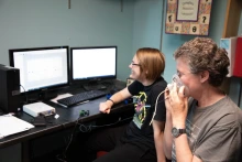 Two people sit in front of computer screens, with one of the individuals holding an oxygen mask to their face.