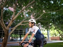 A cyclist wearing a white helmet and blue jersey rides a bike on a tree-lined path next to a building with large windows.
