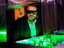A man wearing safety glasses and a suit observes a green-lit optical experiment with lasers and mirrors in a laboratory setting.