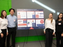 A group of four individuals stands in front of a large poster titled "What Drives Employment Success?" The poster includes charts and bullet points about education, income, and job stress. The group smiles at the camera, wearing business casual attire, with a green and gray wall in the background.