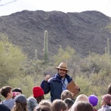 A man in a blue rain coat wearing a woven hat stands in the desert in front of a group of grade school children as he points behind him at the desert landscape.